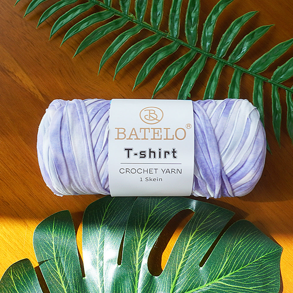 Single lavender-and-white variegated BATELO T-shirt crochet yarn skein on a wooden table with green leaves