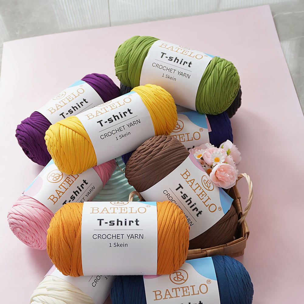 Assorted BATELO T-shirt yarn skeins stacked with flowers on a soft pink tabletop