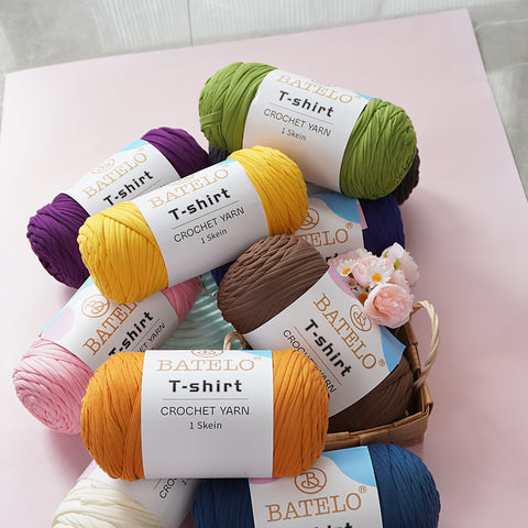 Assorted BATELO T-shirt yarn skeins stacked with flowers on a soft pink tabletop