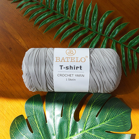 Single light gray BATELO T-shirt crochet yarn skein on a wooden table with green leaves