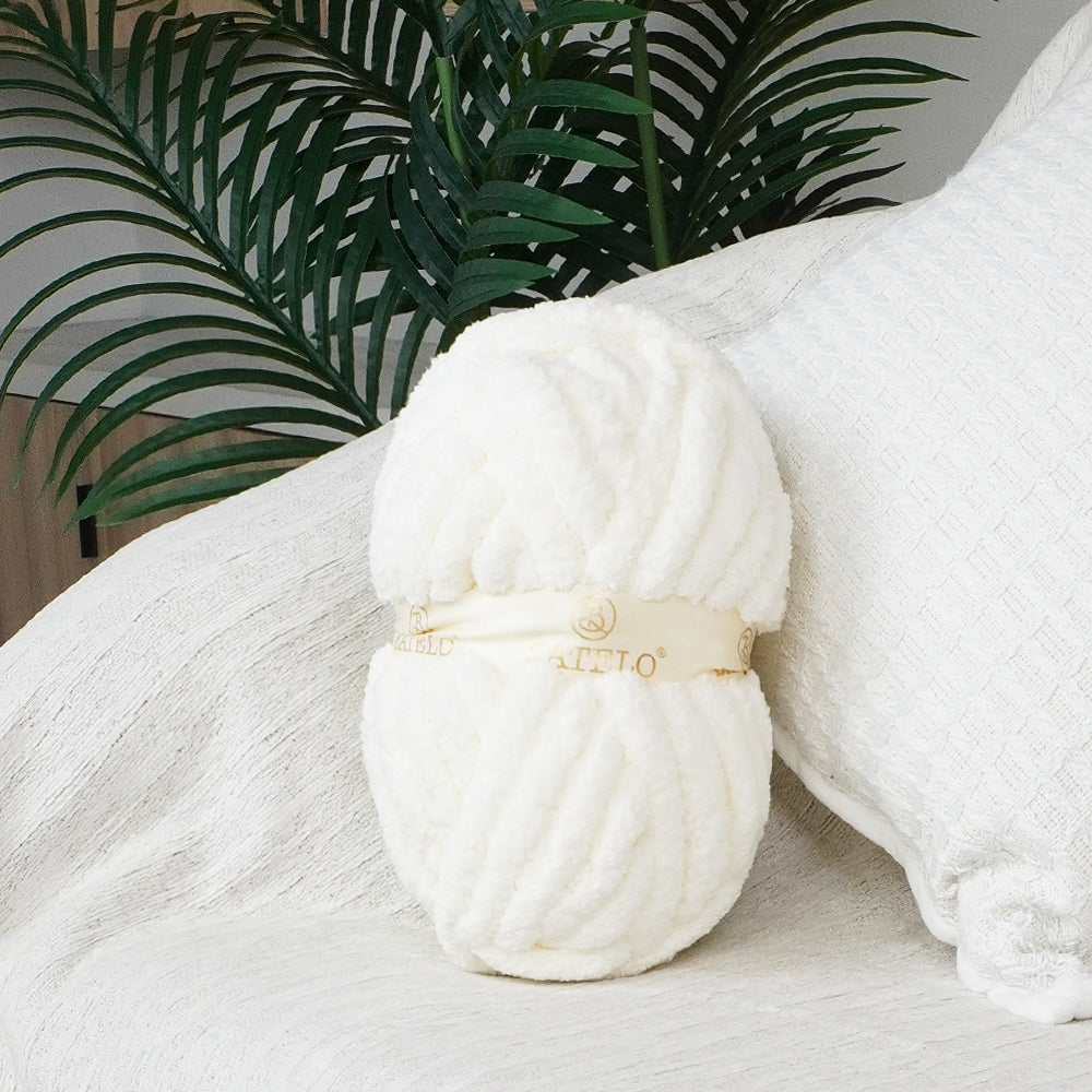 Single white BATELO chunky chenille yarn skein styled on a white sofa with green leaves