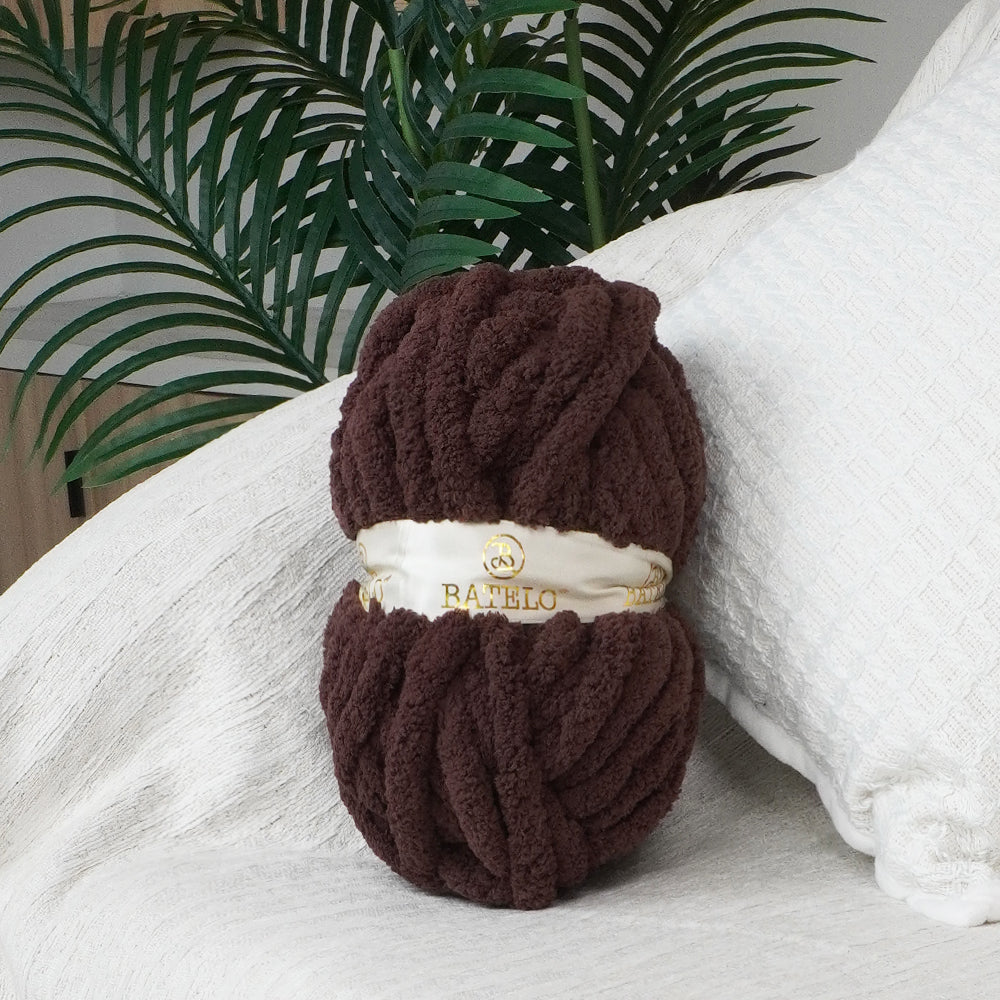 Single dark brown BATELO chunky chenille yarn skein styled on a white sofa with green leaves