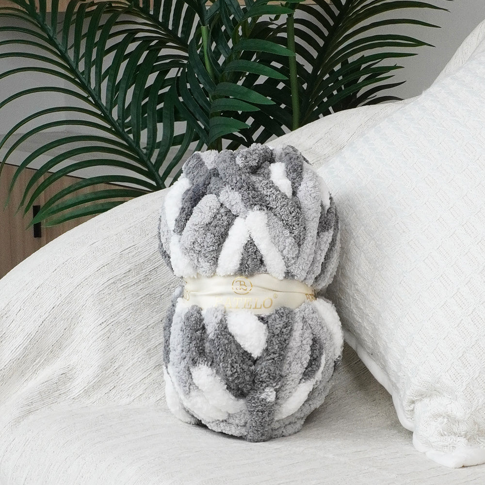 Single gray-and-white variegated BATELO chunky chenille yarn skein styled on a white sofa with green leaves