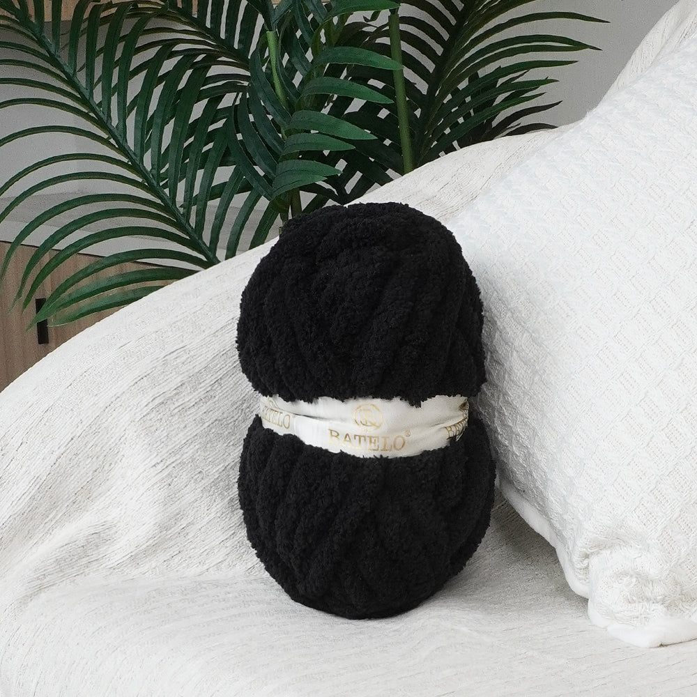 Single black BATELO chunky chenille yarn skein styled on a white sofa with green leaves