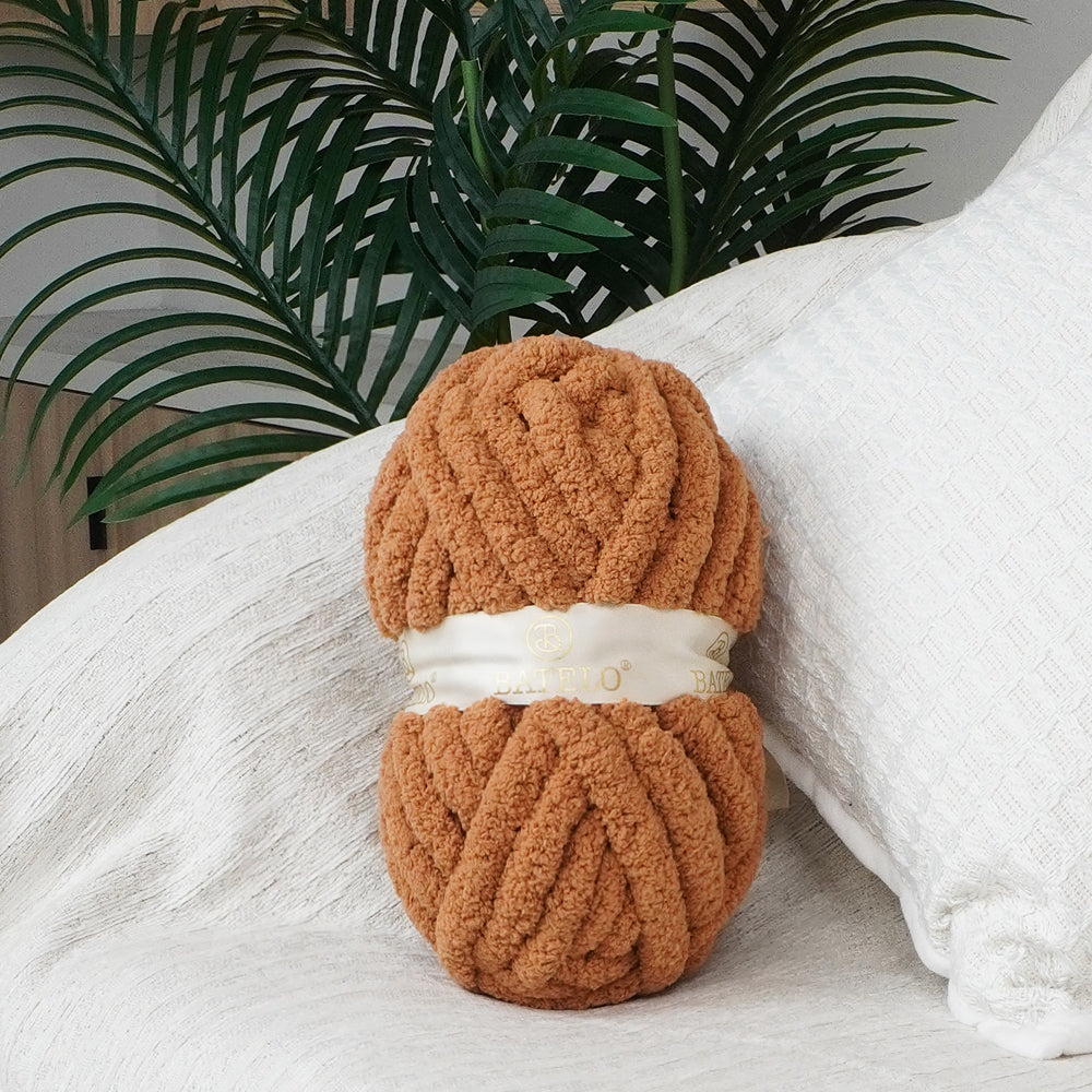 Single camel BATELO chunky chenille yarn skein styled on a white sofa with green leaves