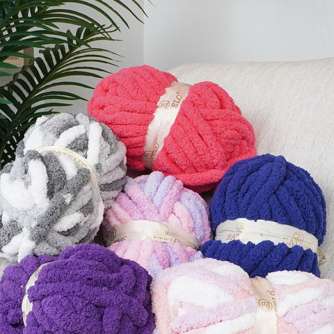 Assorted BATELO chunky chenille yarn skeins in pink, purple, blue, and gray piled on a white sofa