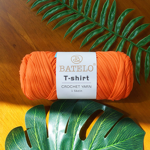 Single orange BATELO T-shirt crochet yarn skein on a wooden table with green leaves
