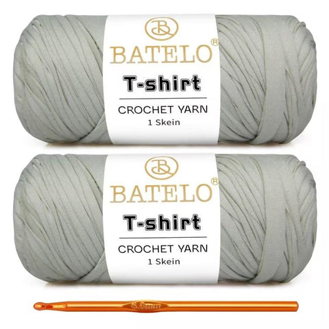 Two pale gray BATELO T-shirt yarn skeins with a gold crochet hook on a white background