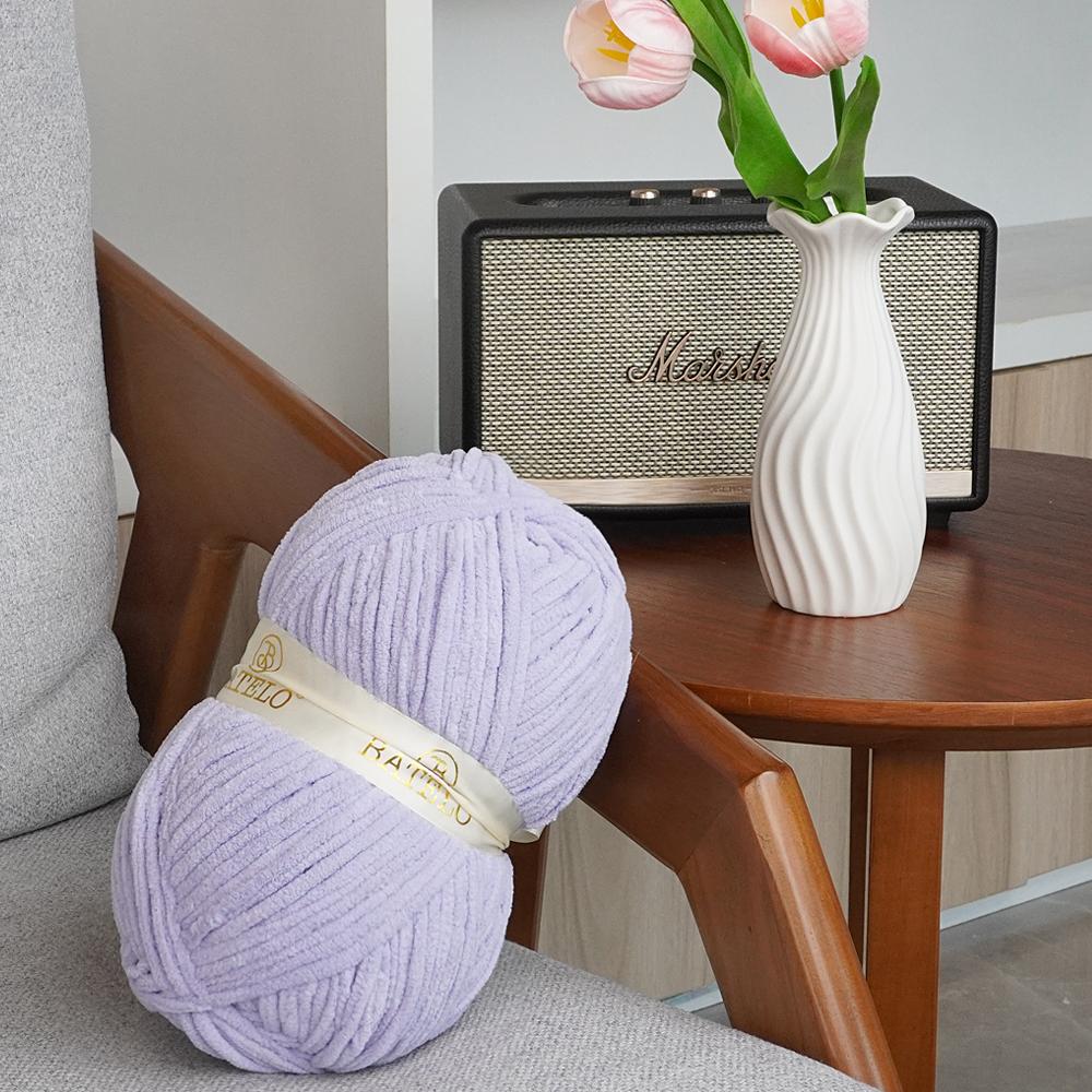 Single lavender BATELO bulky chenille yarn skein on a chair beside flowers and a speaker