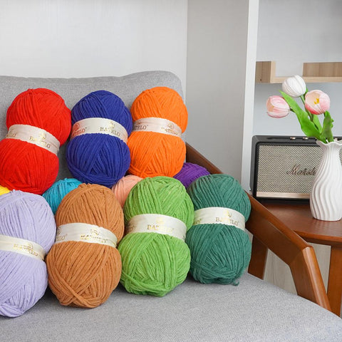Assorted BATELO bulky chenille yarn skeins in vivid colors on a gray chair beside flowers and a speaker