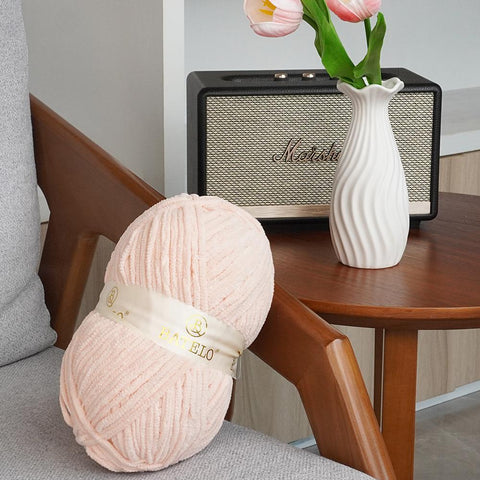 Single pale pink BATELO bulky chenille yarn skein on a chair beside flowers and a speaker