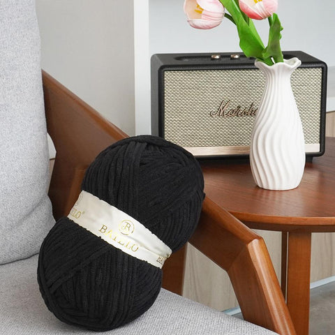 Single black BATELO bulky chenille yarn skein on a chair beside flowers and a speaker